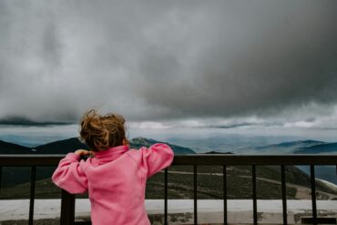 girl standing near railing with mountains and clouds in the distance